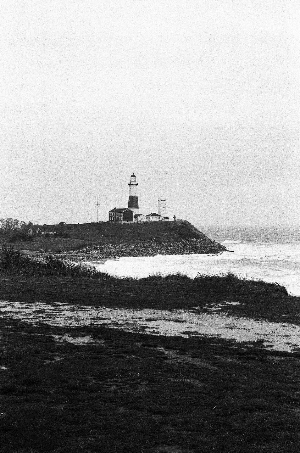 A lighthouse stands on a hill overlooking a rough sea with waves crashing against the shore.