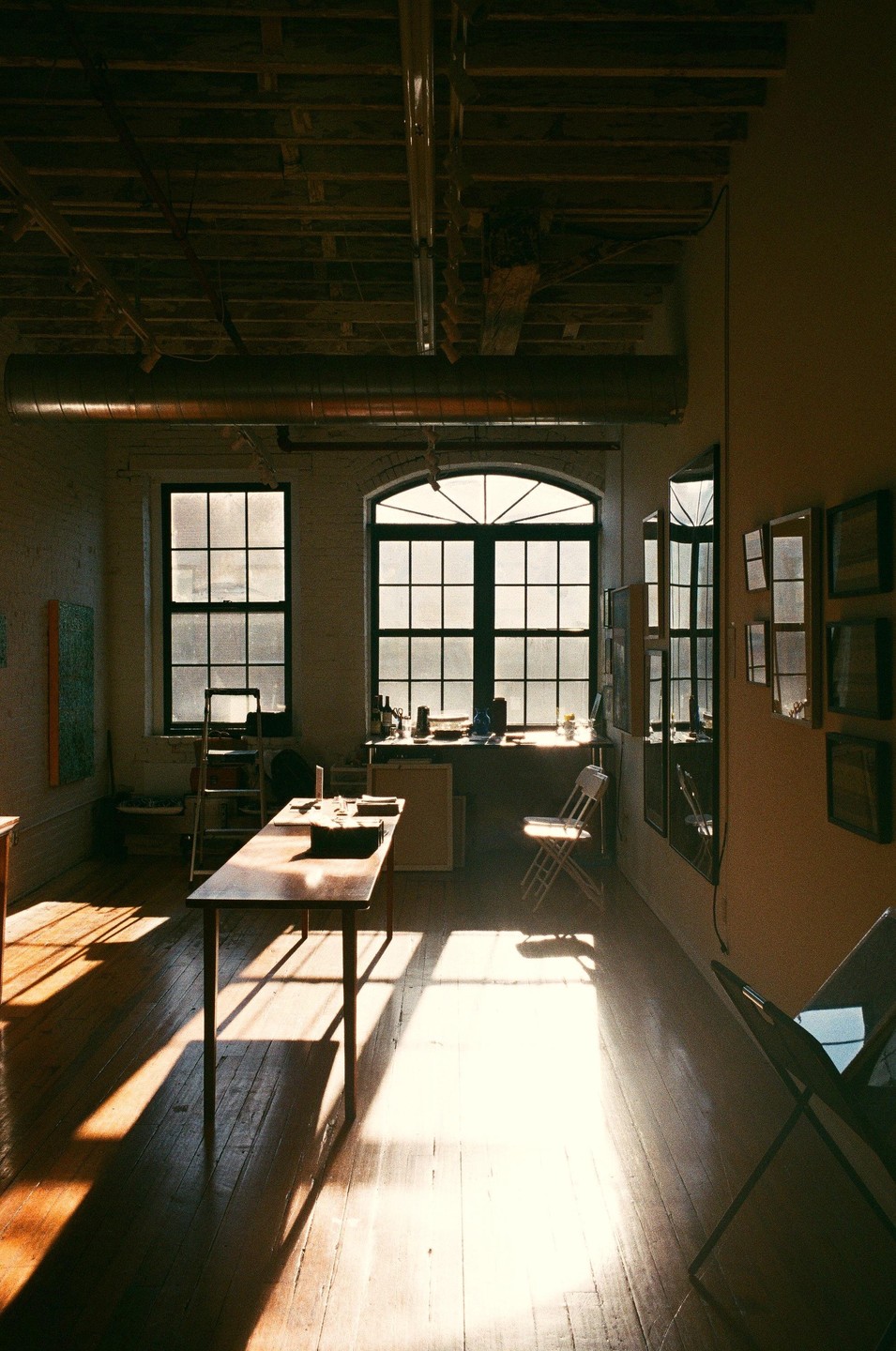 Sunlight streams through large windows into an artist's studio with a long table and chairs.