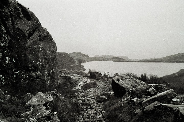 A rocky path leads to a lake surrounded by hills under a misty sky.