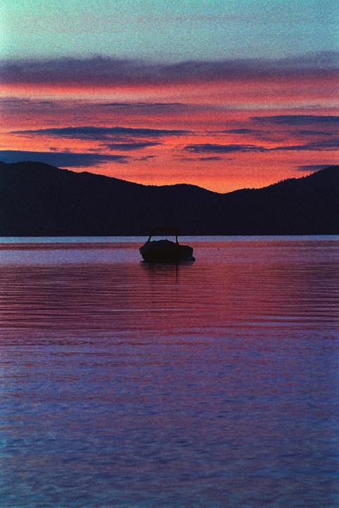 A boat floats on a lake with mountains in the background under a colorful sky at sunrise.
