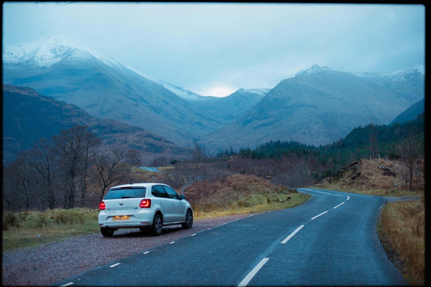 A white car is parked on the side of a winding road with snow-capped mountains in the background.