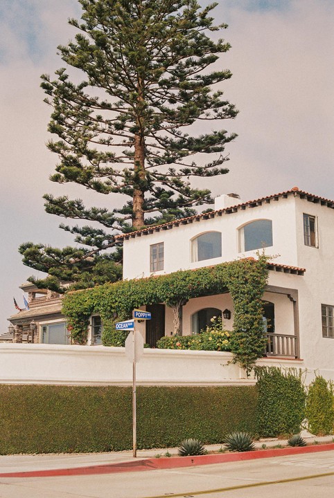 A tall pine tree stands next to a white stucco house with a red tile roof and green vines covering its facade, with a street sign for Poppy Avenue and Ocean Boulevard in the foreground.
