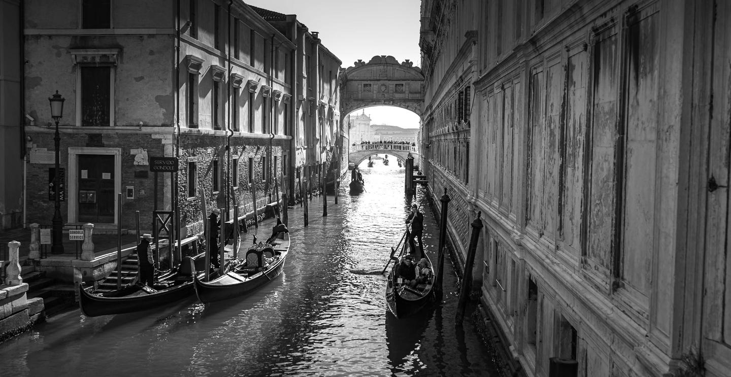 Gondolas are visible on a canal in Venice, with historic buildings lining both sides and a bridge in the distance.