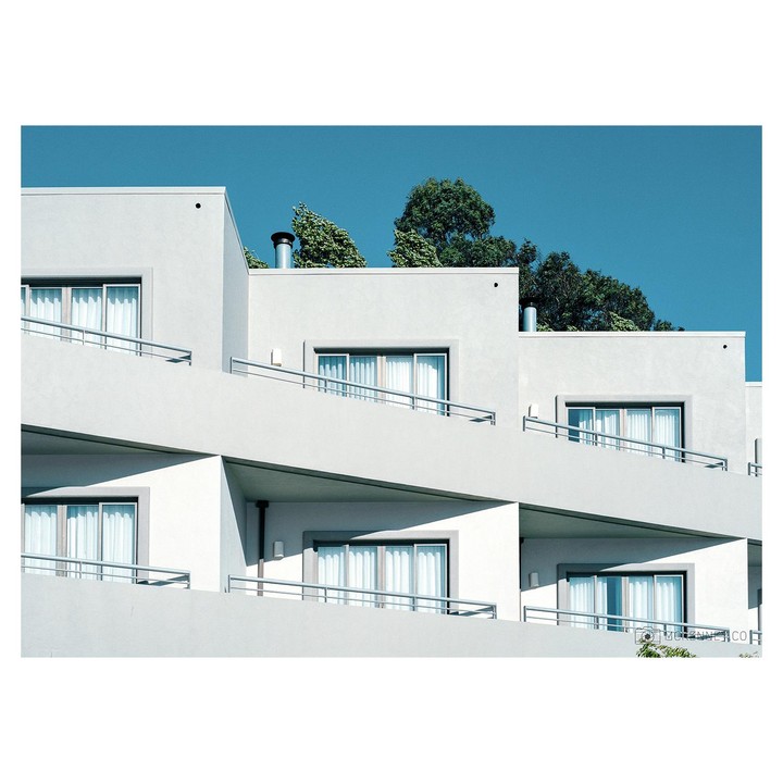 A white building with multiple balconies and windows is shown against a clear blue sky with green trees visible above the roofline.