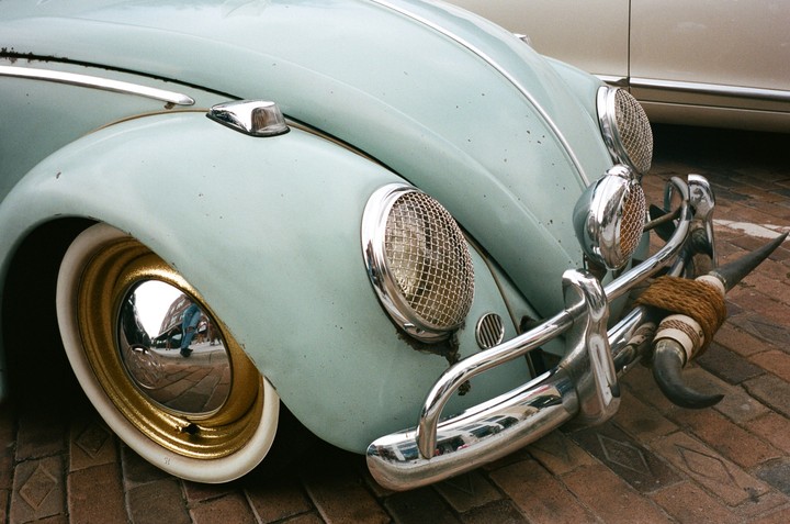 A light blue vintage car with bull horns attached to its front bumper is parked on a brick surface.