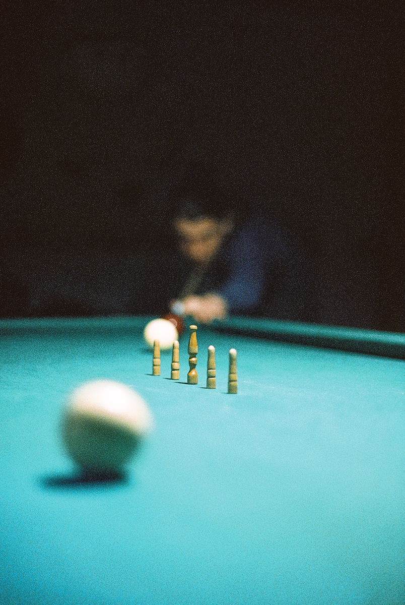 A man plays billiards with a cue ball in the foreground and wooden pins on the table.