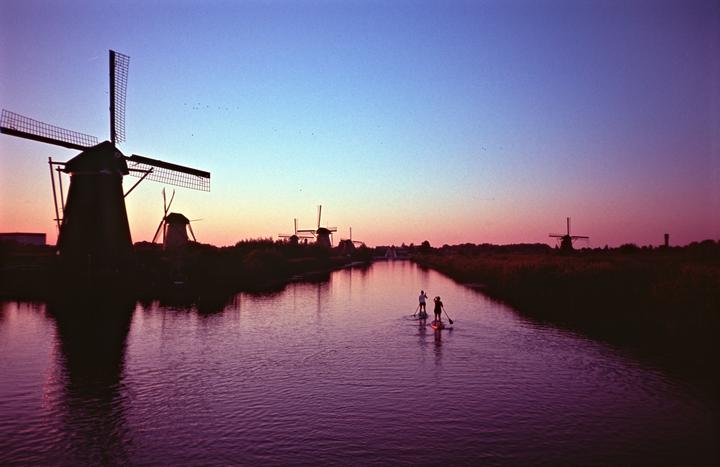 Two people paddleboard on a canal lined with windmills at dusk.