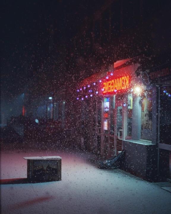 Snow falls in front of a storefront with a neon sign and string lights.