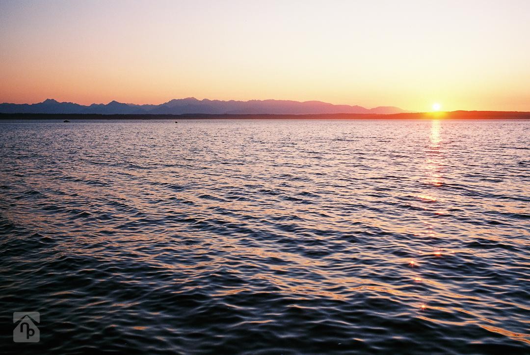 The sun sets over a body of water with mountains in the distance.
