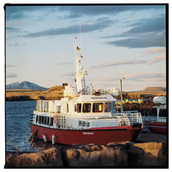 A red and white boat named Skrúður is docked in a harbor with mountains in the background under a cloudy sky.