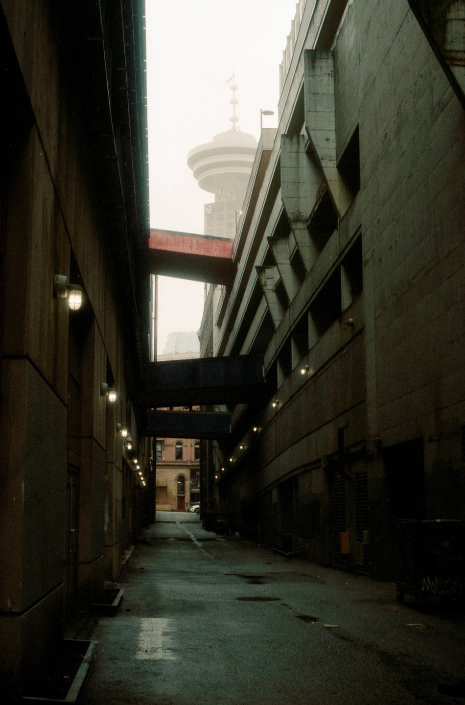 A narrow alleyway with concrete buildings on either side leads to a distant tower visible through a gap.