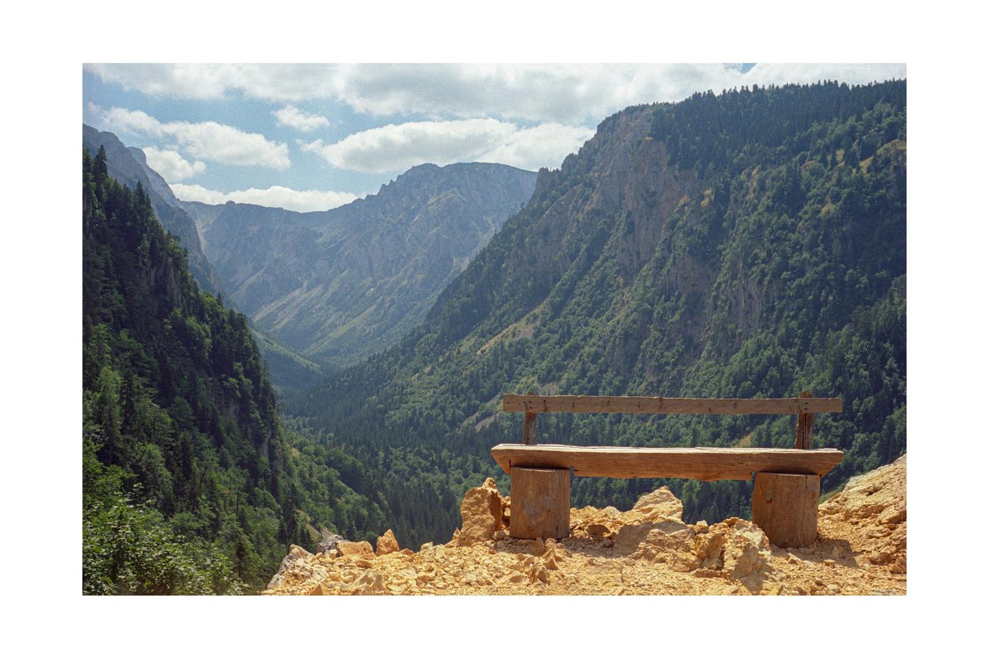 A wooden bench sits on a rocky outcrop overlooking a deep valley with forested mountains under a cloudy sky.