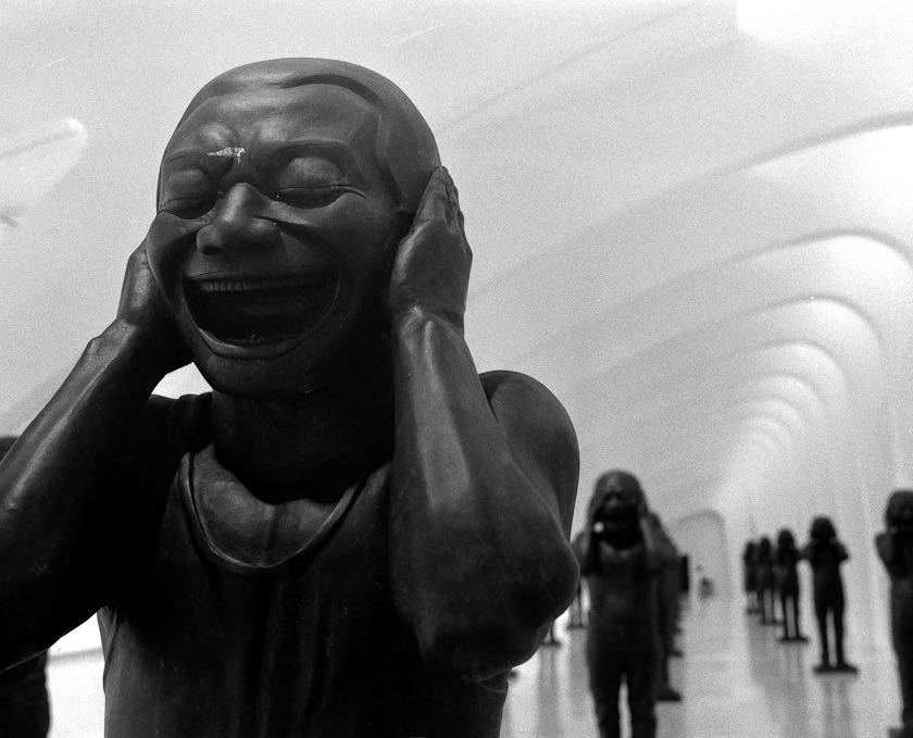 A statue with a wide open mouth and hands covering its ears stands in a long hallway lined with similar statues.