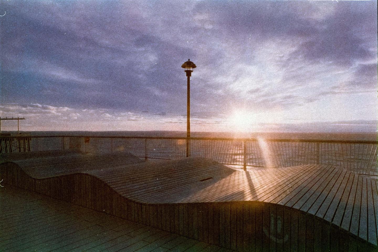 The sun sets over the ocean, casting a warm glow on a wooden boardwalk with a single lamppost.