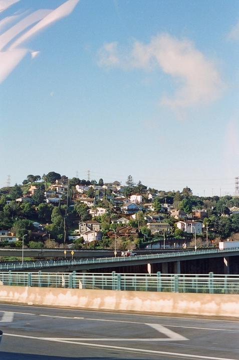 A hillside neighborhood with many houses and trees overlooks a highway and bridge.