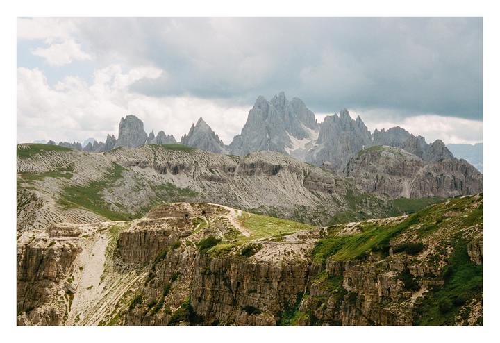 A hiking trail winds through a mountainous landscape with jagged peaks under a cloudy sky.