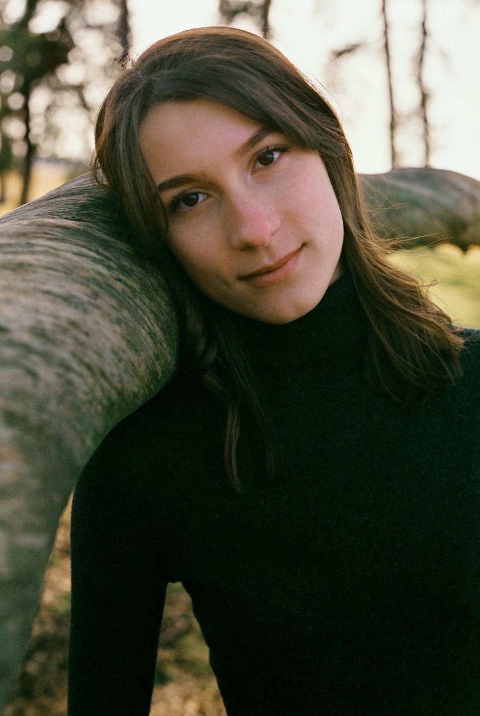 A woman with long hair and a dark top leans her head on a tree trunk.