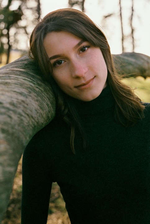 A woman with long hair and a dark top leans her head on a tree trunk.