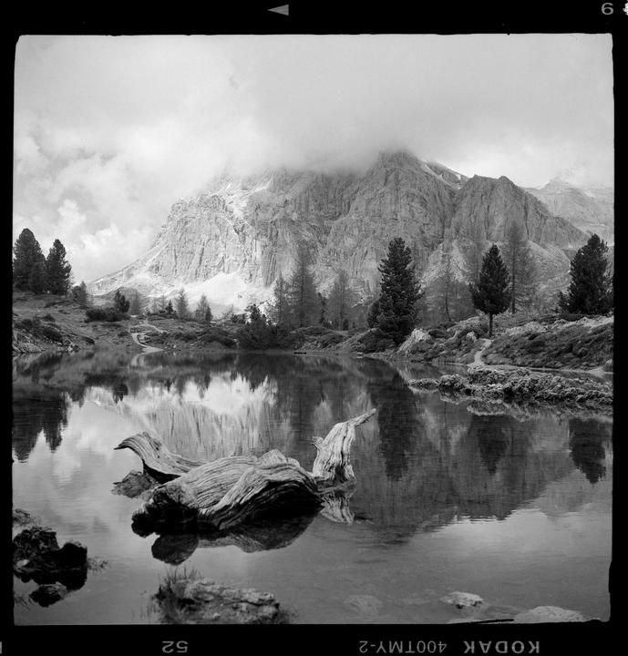 A mountain with clouds at its peak is reflected in a lake with a piece of driftwood in the foreground.