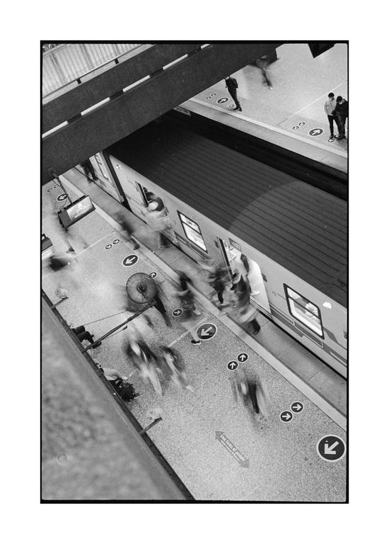 A high-angle, black and white shot shows a train station platform with a train and people with motion blur.