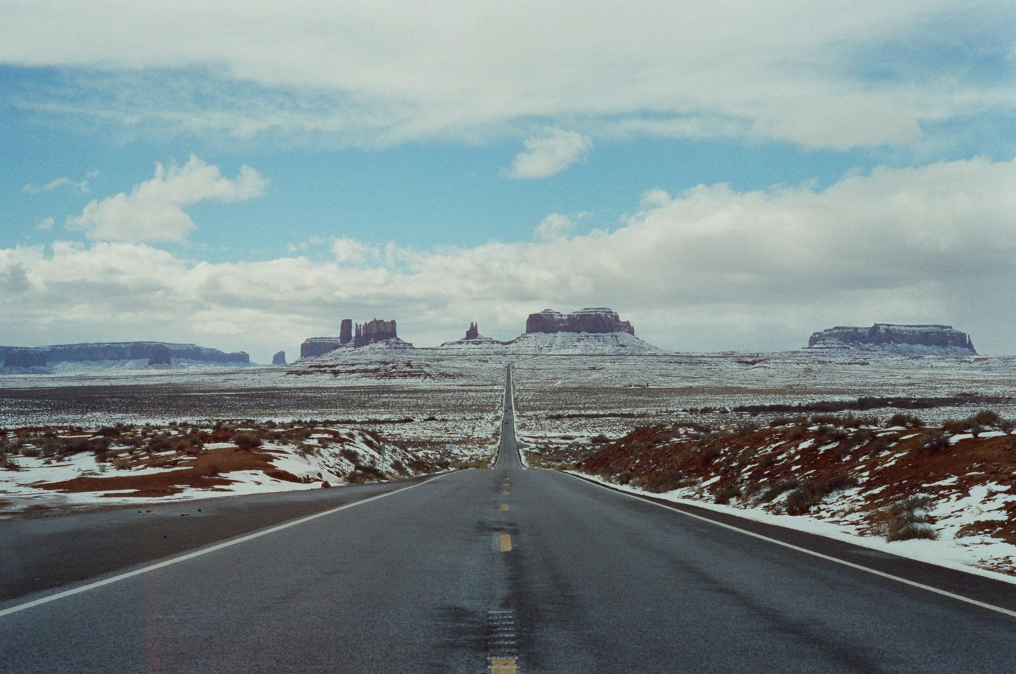 A long, straight road leads towards distant mesas and buttes in a snow-dusted desert landscape under a cloudy sky.