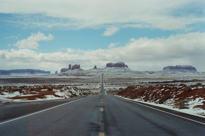 A long, straight road leads towards distant mesas and buttes in a snow-dusted desert landscape under a cloudy sky.