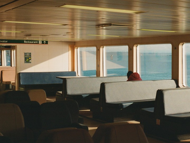 A person in a red hoodie sits on a ferry looking out at the water through large windows.