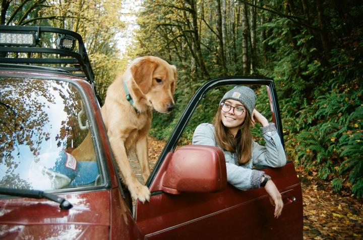 A woman and a dog are in a red vehicle on a dirt road surrounded by trees.