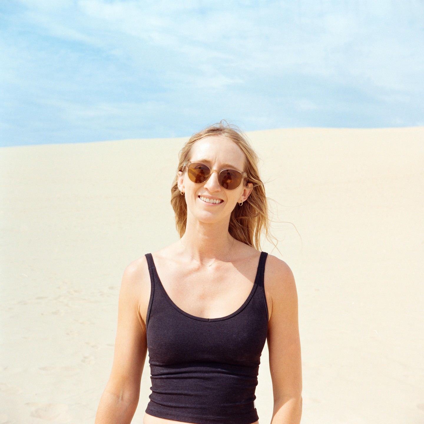 A woman wearing sunglasses and a black tank top smiles at the camera with a sand dune and blue sky in the background.
