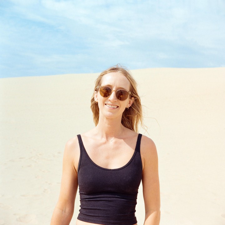 A woman wearing sunglasses and a black tank top smiles at the camera with a sand dune and blue sky in the background.