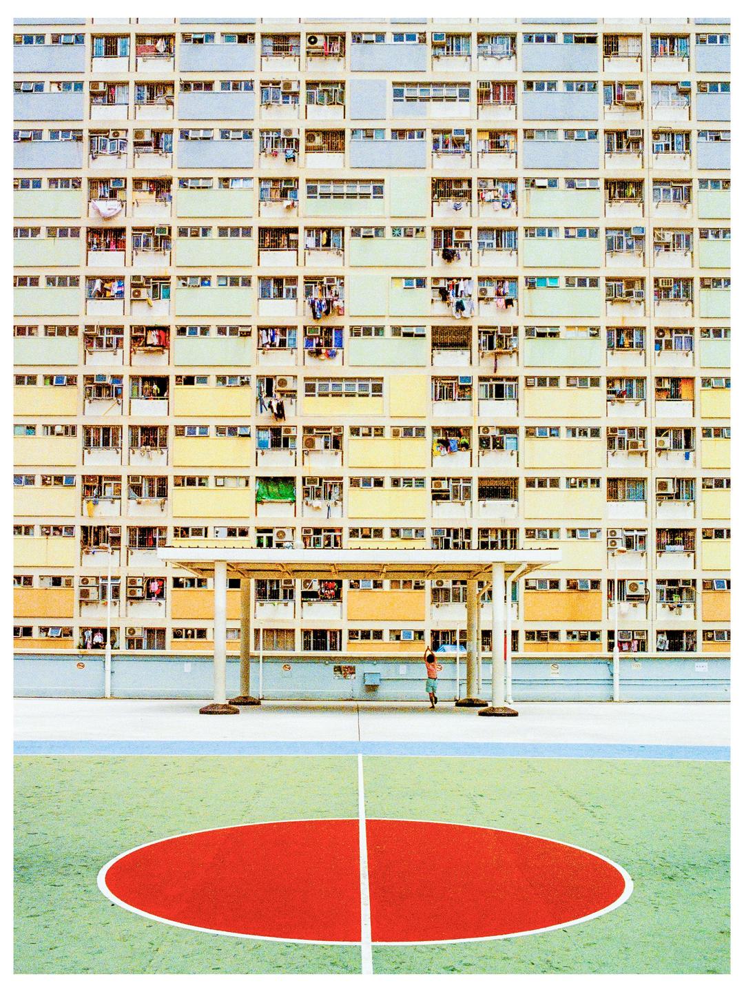 A child stands on a basketball court in front of a large apartment building in Hong Kong.