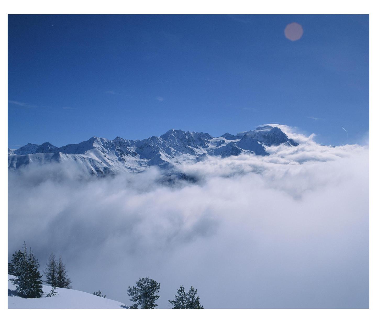 Snow-covered mountains rise above a sea of clouds under a clear blue sky.