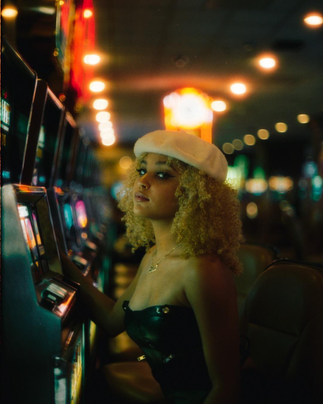 A woman with curly hair and a white beret sits in front of a row of slot machines.