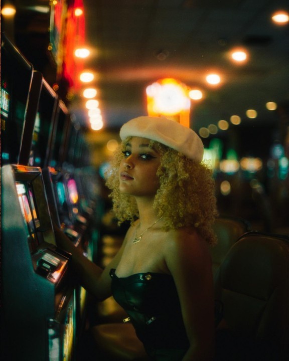 A woman with curly hair and a white beret sits in front of a row of slot machines.