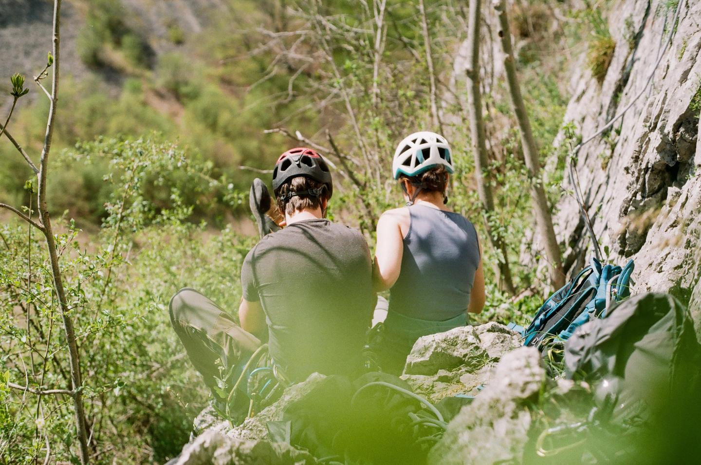 A man and a woman wearing helmets sit on a rocky outcrop with climbing gear next to a cliff face.