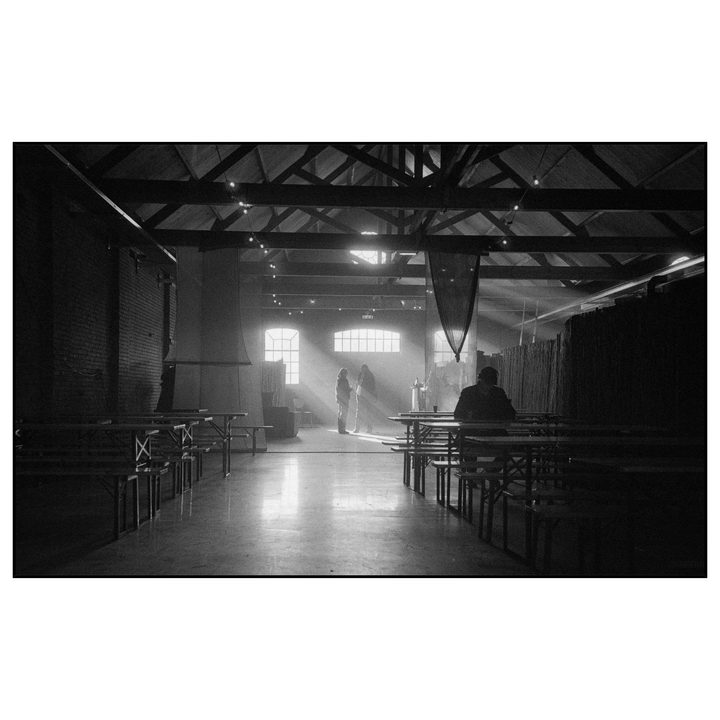 Sunlight streams through windows into a large, empty hall with tables and benches, illuminating two people standing in the distance and one man seated in the foreground.