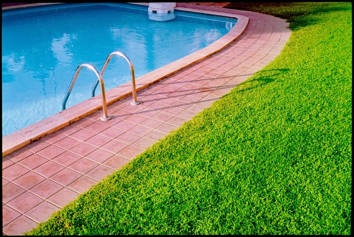 A swimming pool with a metal ladder is next to a tiled path and a patch of green grass.