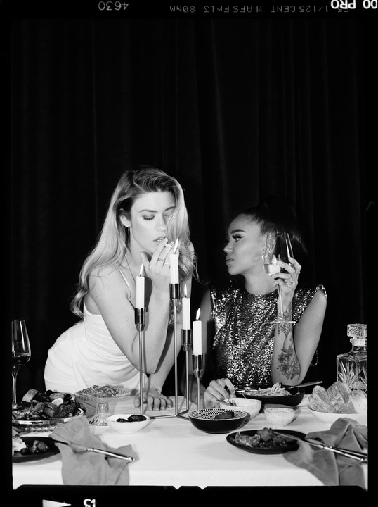 Two women sit at a table with lit candles and food, one smoking a cigarette and the other holding a glass.