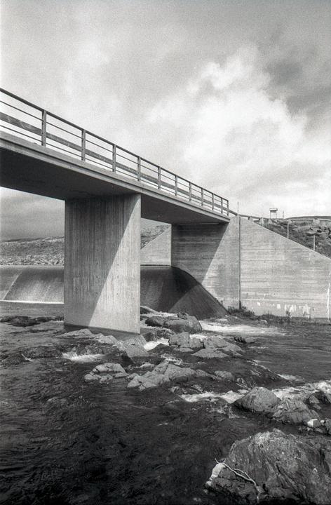 A concrete bridge with a metal railing spans over a river with rocks and a small waterfall.