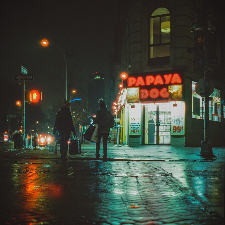 Two people walk away from a Papaya Dog restaurant on a wet street at night.