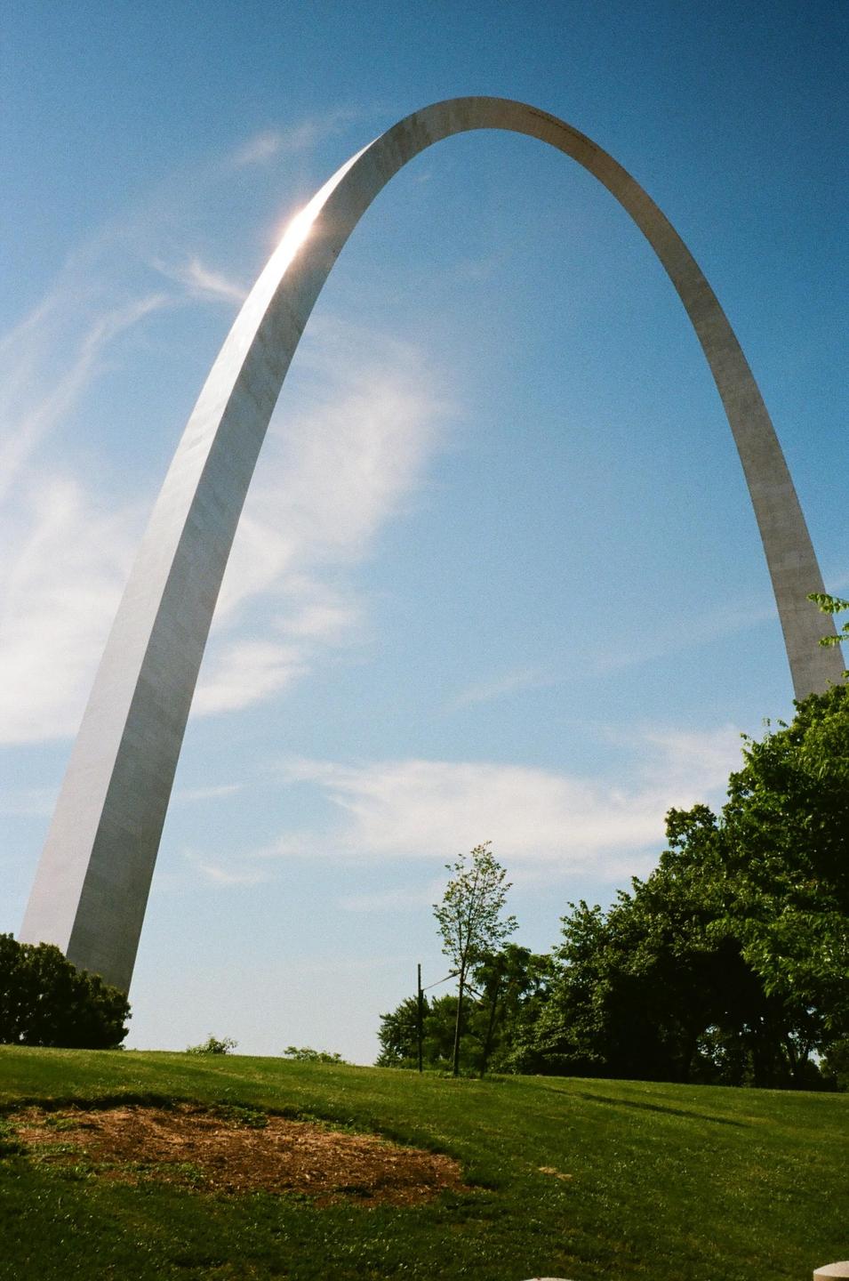 The Gateway Arch stands tall against a blue sky with a sun flare on its side.