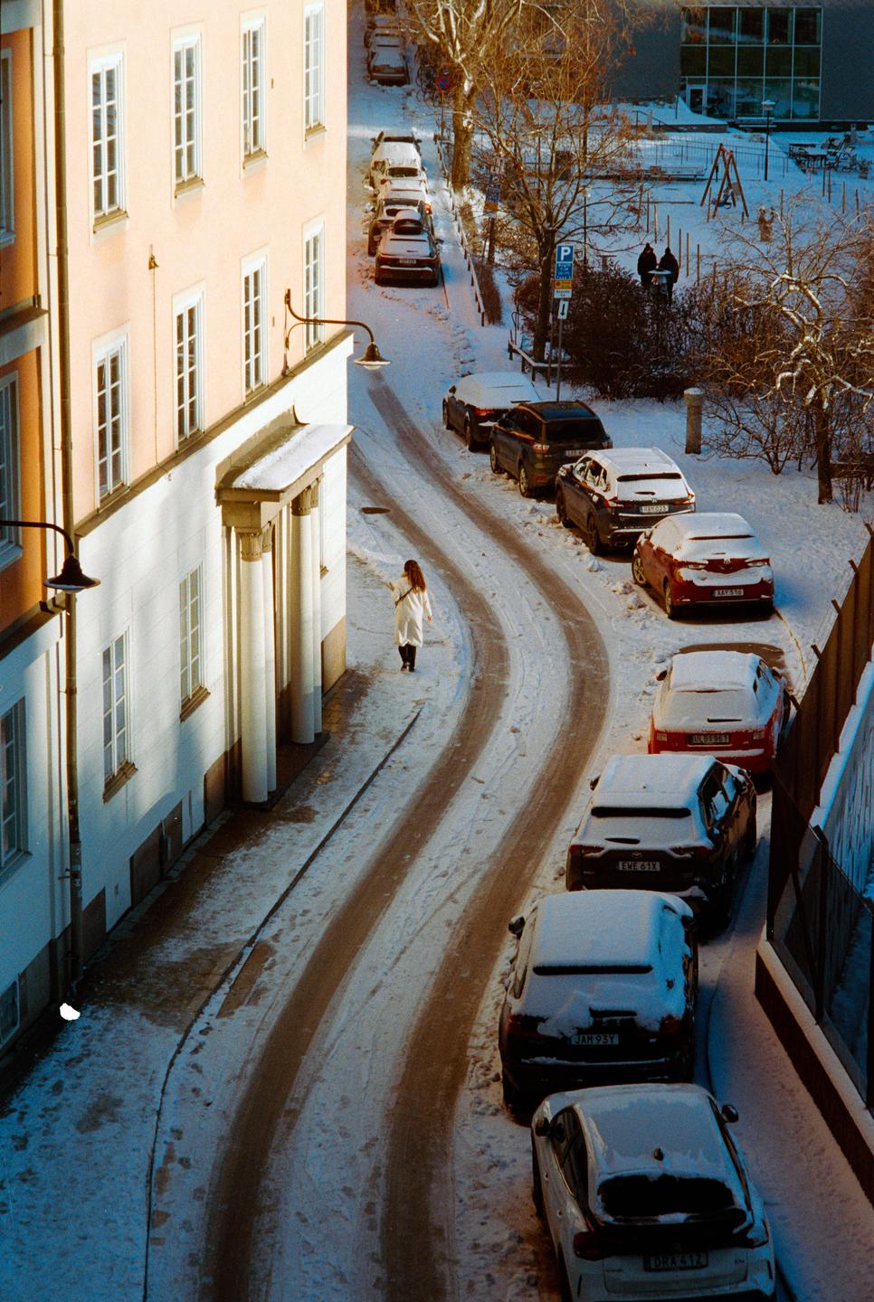 A woman walks along a snow-covered street lined with parked cars and buildings.