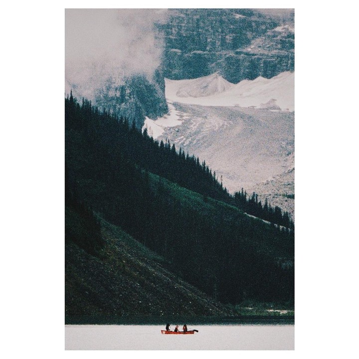 A red canoe with three people floats on a lake surrounded by a forest and snowy mountains.