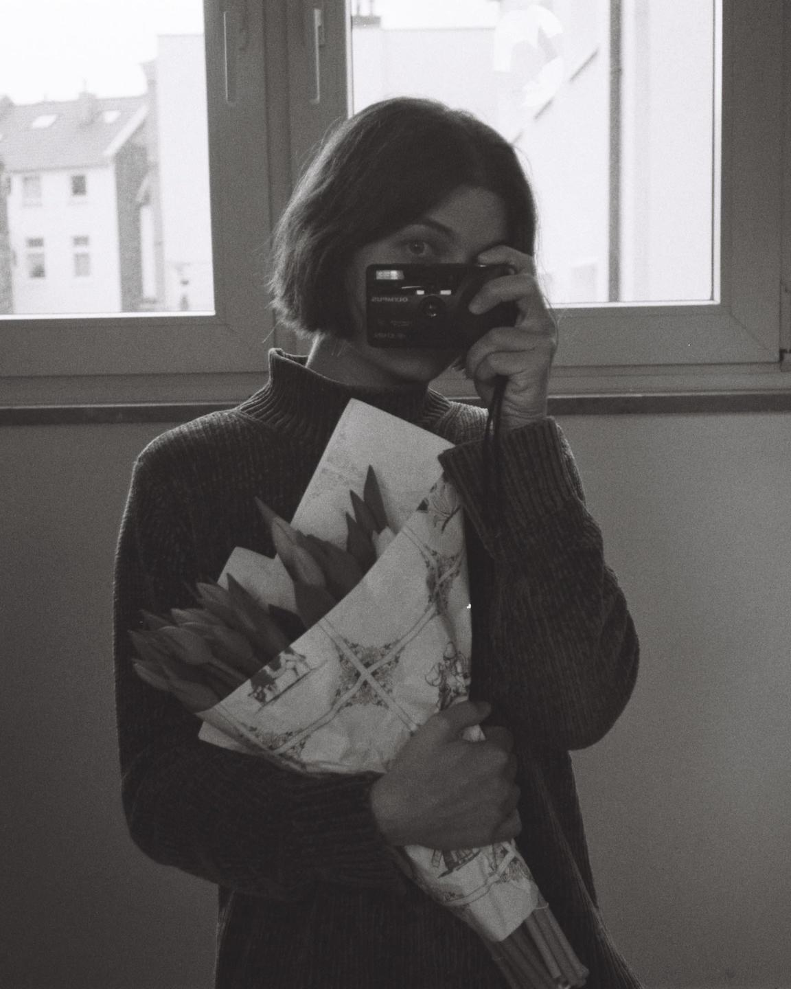 A woman holds a bouquet of tulips wrapped in patterned paper while holding a camera to her eye.