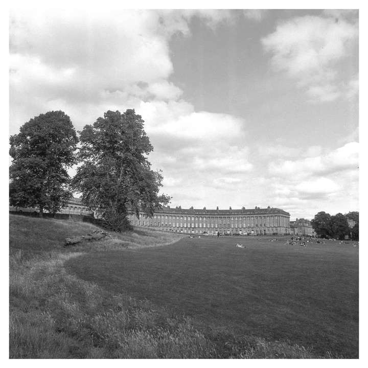A large curved building with many windows stands behind a wide grassy field where people are relaxing.