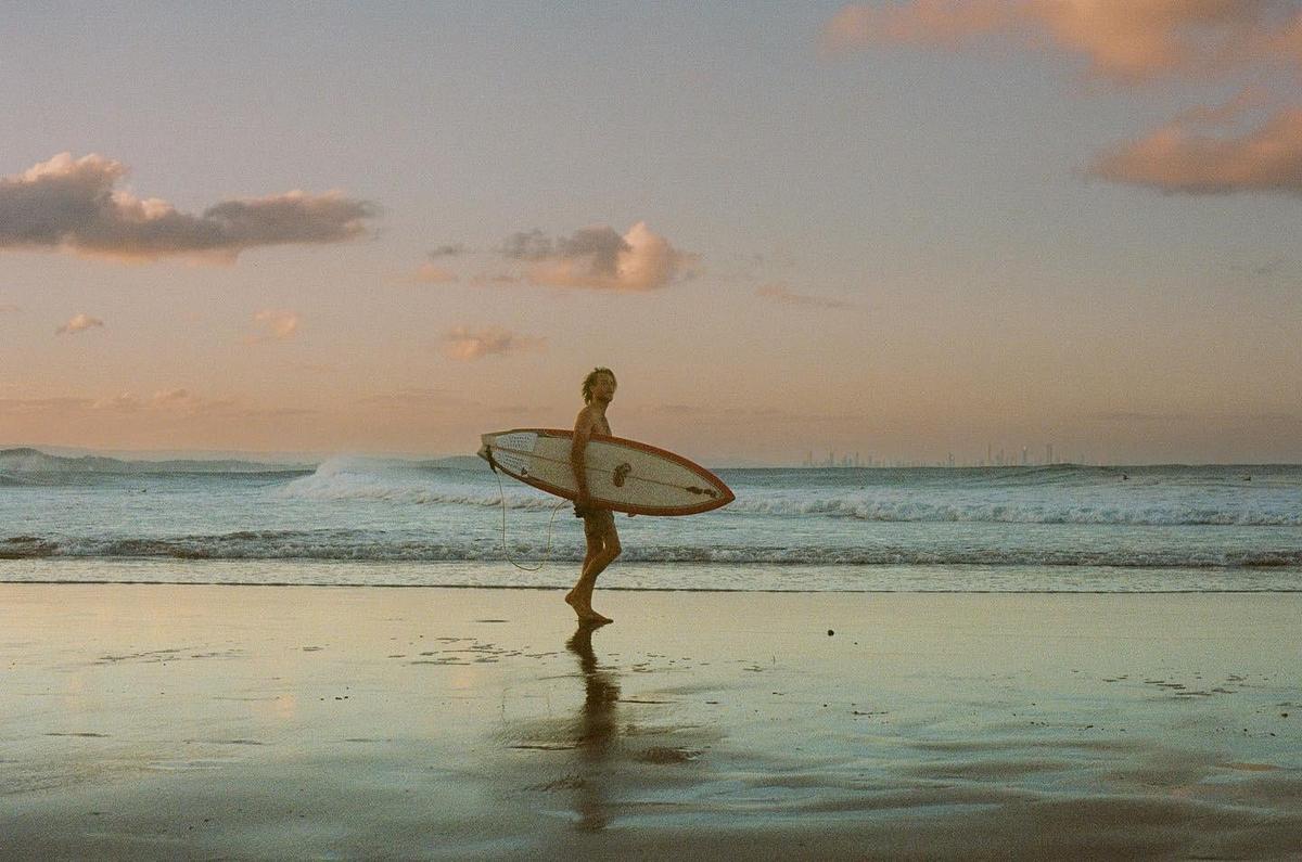 A man carries a surfboard on a beach at sunset with waves crashing in the background.