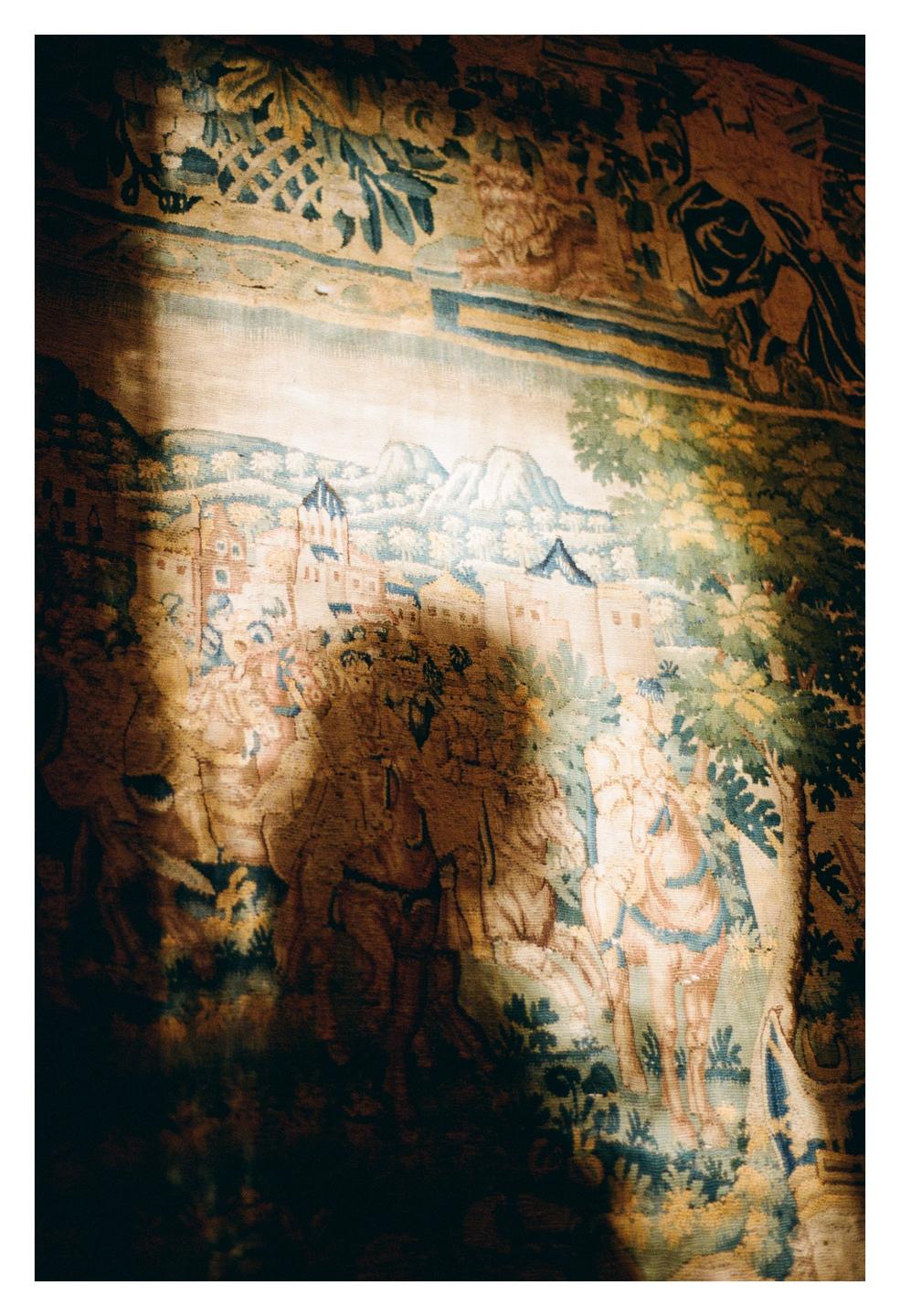 A tapestry depicting a castle, mountains, and people on horseback is illuminated by a shaft of light.