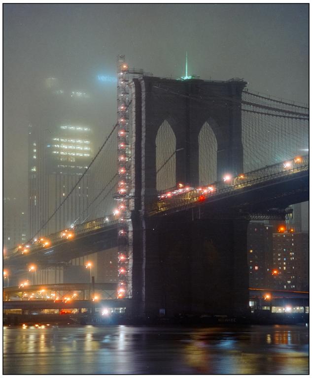 The Brooklyn Bridge is illuminated at night, with traffic visible on its roadway and city lights reflecting on the water below.