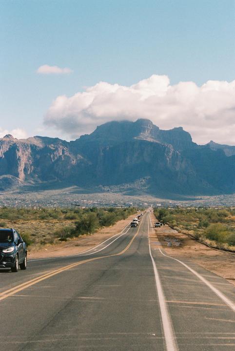 A long road leads toward a large mountain range under a cloudy sky.
