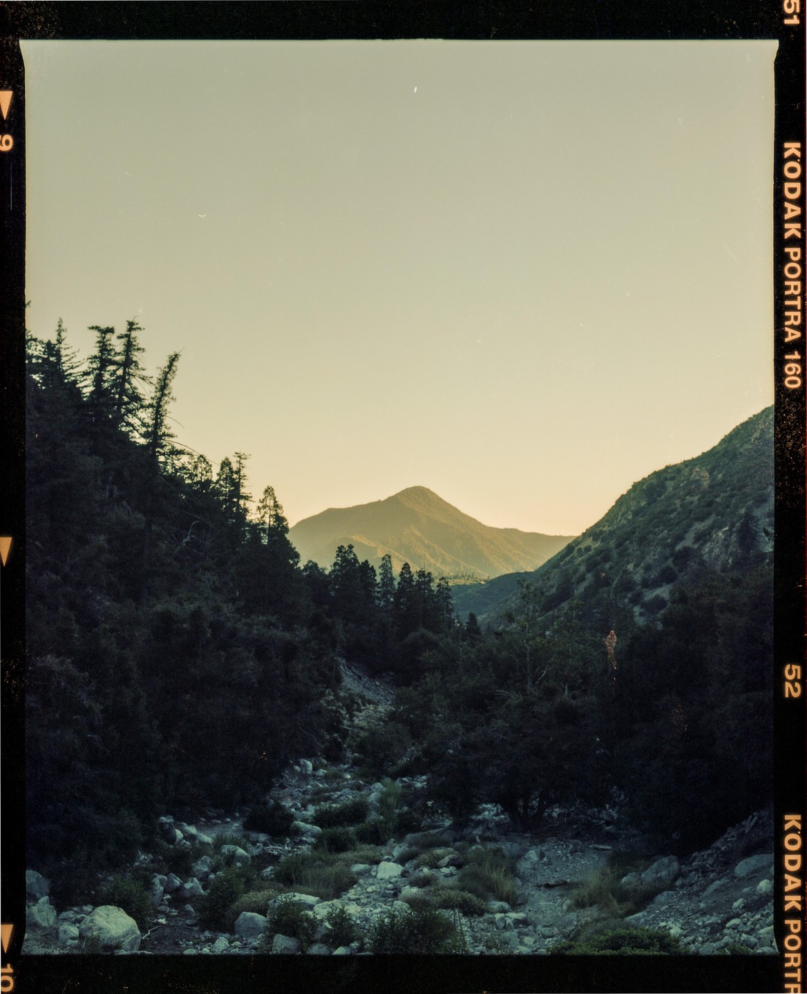 A mountain valley with a dry riverbed is surrounded by dark trees and mountains under a pale sky.